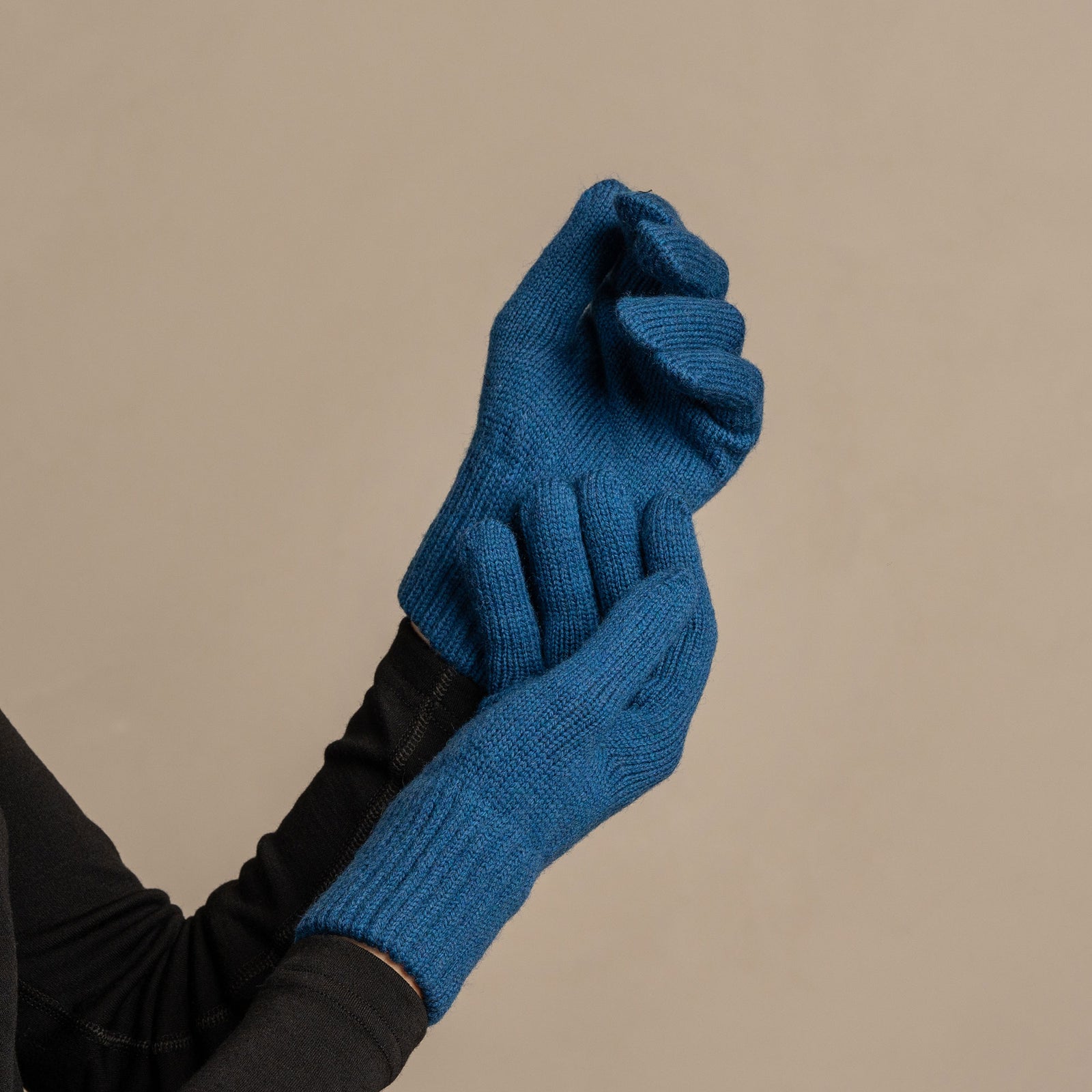 A close-up shot of a person wearing menique brand vibrant blue, ribbed knit gloves. The person is wearing a dark long-sleeved top, with the right hand holding the left wrist, showcasing the gloves' fit and the small, rectangular tan leather menique tag on the cuff of the left glove. The background is a solid, neutral gray.