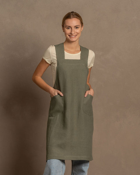 A fair-skinned woman with blonde hair pulled back smiles at the camera. She is wearing a white short-sleeved shirt under a stone green linen cross-back apron with pockets, and light blue jeans are visible below the apron. She stand against a plain warm brown background.