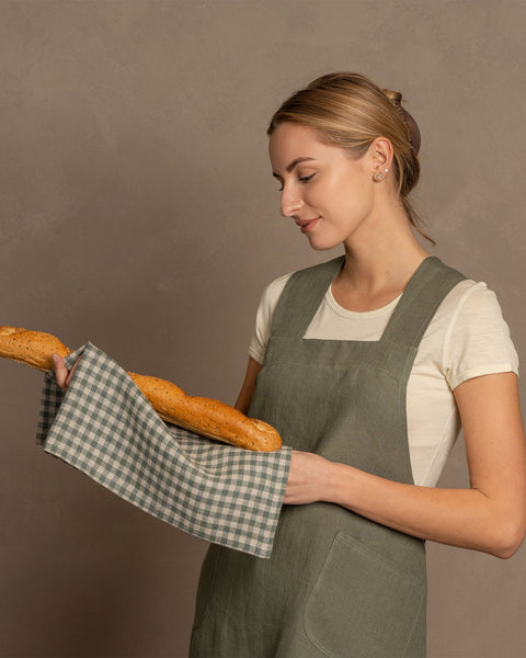A woman wearing a natural white t-shirt under a sage green, cross-back linen apron. She is looking down with a slight smile while holding a long, seeded baguette wrapped in a green and white checkered linen tea towel. The background is a solid, warm taupe wall.