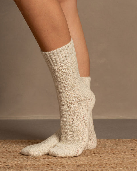 A low-angle shot shows a person&#39;s legs and feet, likely wearing menique brand off-white, crew-length knitted wool socks with a visible ribbed cuff and textured cable-knit pattern on the foot. The person is standing on a light brown, woven mat, against a neutral, plain background.