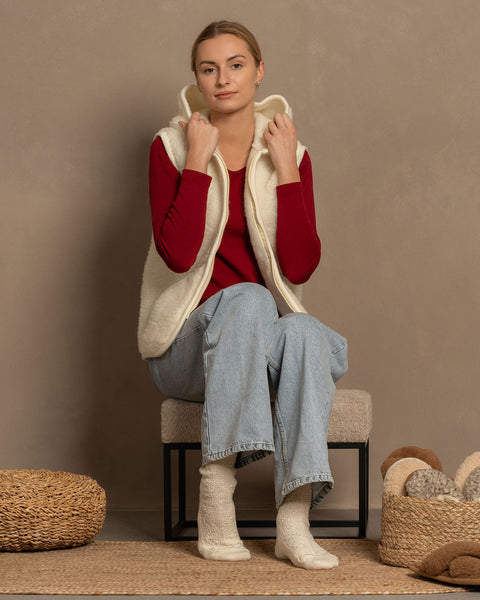 
A light-skinned person with blonde hair is seated on a stool, wearing a red long-sleeved shirt, light-wash blue jeans, menique brand off-white knitted socks, and a cream-colored hooded fleece vest. Their hands are holding the hood of the vest. To the left is a round woven pouf, and to the right, a woven basket contains several pairs of slippers. They are all positioned on a woven mat against a neutral wall.