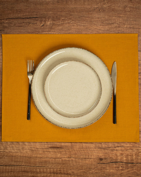 The menique linen placemat in spicy yellow color. There are two porcelain plates in different sizes on top of it, and cutlery by the sides of them. There is a wooden tabletop visible in the background.