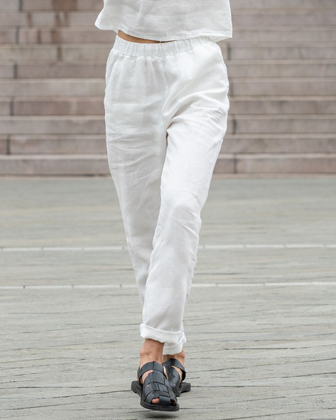 A low-angle, full shot shows a person from the waist down, walking forward on a paved surface, wearing pure white linen menique pants and dark sandals. The pants are slightly wrinkled, giving them a relaxed look, and are cuffed at the ankles.