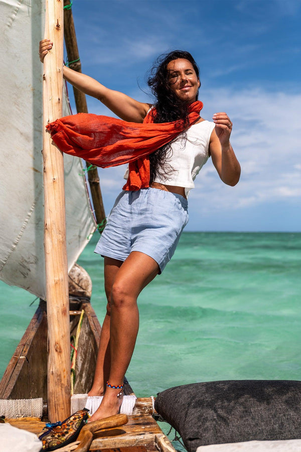 A woman stands on a rustic wooden boat with a white sail against a bright blue sky and turquoise water. She wears a white sleeveless top, light blue shorts, and an orange scarf. The boat has a white cushion and a dark gray cushion.