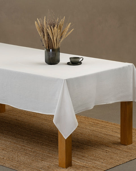 The menique linen tablecloth in pure white color. The tablecloth drapes over a wooden table, there is a vase and a cup on top of it.