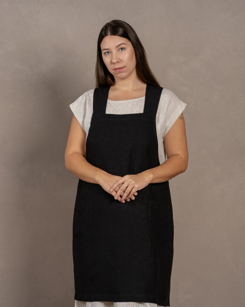 A young woman with dark hair standing in a studio background, with her hands put together in front. She is wearing a linen dress with a linen cross-back japanese apron in pure black color in size L/XL on top.