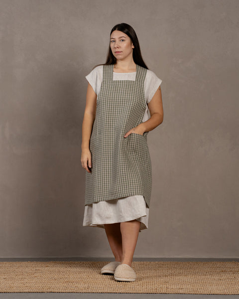 A woman with long dark hair stands facing the camera, wearing a linen menique cooking apron over a white, patterned dress. She also wears light-colored slippers and stands on a woven rug against a mottled gray background.