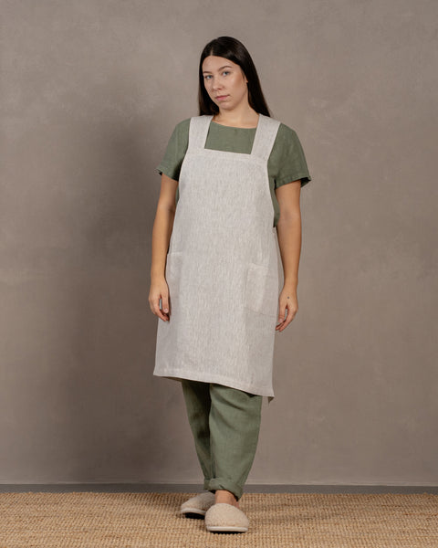 A woman wearing a natural color linen apron over a stone green outfit, standing against a textured background.