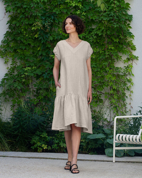 A woman with short, dark, curly hair walks on a light-colored stone surface, looking towards the left. She is wearing a menique linen, v-neck dress with short, slightly ruffled sleeves and a gathered, tiered hem. Her hands are casually placed in the side pockets of the dress. She is wearing black, open-toed sandals. Behind her is a light-colored wall partially covered with lush green climbing plants. 