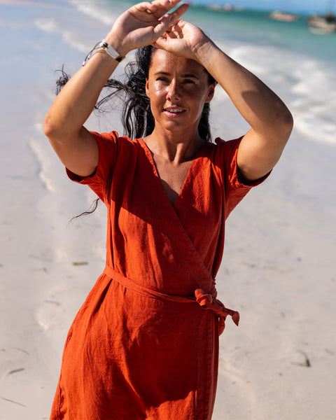 A smiling woman with dark hair stands barefoot on a sandy beach, wearing a cinnamon red wrap dress with short sleeves. She has both arms raised above her head with open hands. The ocean is in the background with gentle waves and several boats visible under a bright sky.
