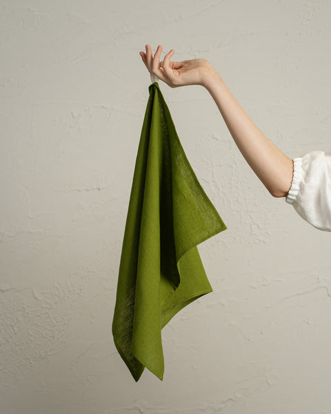A person’s hand holding up a green linen kitchen towel against a textured light wall.