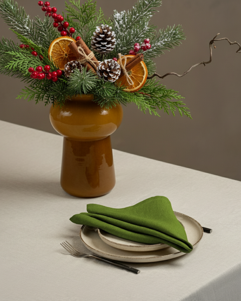 A minimalist holiday table setting featuring a folded forest green napkin placed on stacked ceramic plates beside a fork, with a brown vase holding festive greenery, pinecones, red berries, and dried orange slices.