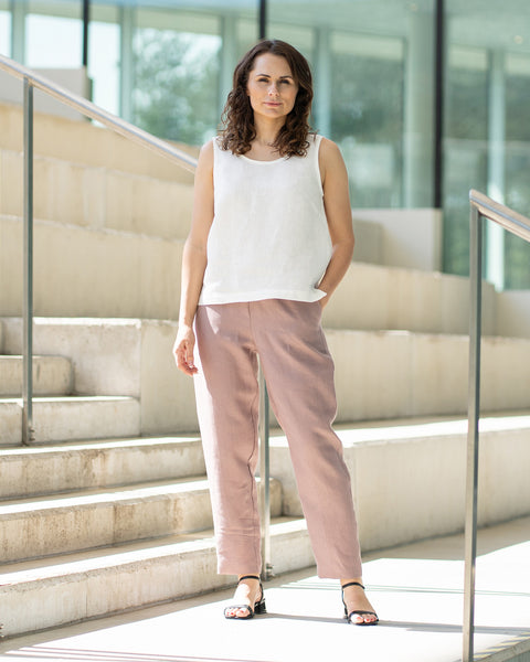 A woman with brown, shoulder-length hair, wearing a white sleeveless top and faded rose trousers, stands on concrete stairs inside a building with glass walls.