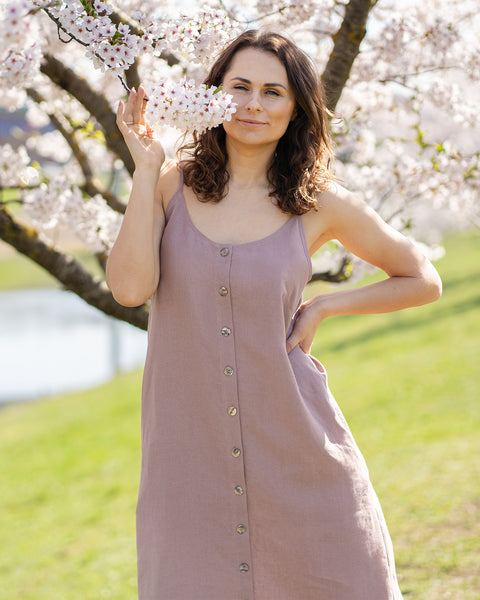 A woman with long, dark, wavy hair, wearing a sleeveless, button-front, faded rose midi-dress, stands outdoors in front of a tree with white blossoms. She holds a sprig of the blossoms up to her nose with her right hand. Her left hand is on her hip. She looks directly at the viewer with a slight smile. The background is a sunny, outdoor setting with green grass and more blossoming trees.