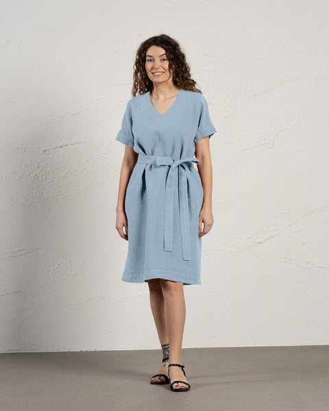 A woman with curly brown hair smiles while modeling a cloudy blue linen menique dress with short sleeves and pockets.