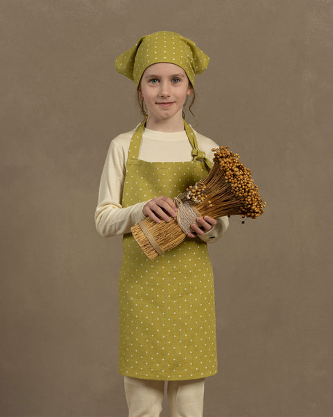 A child stands facing forward, wearing a long-sleeved cream-colored shirt, light-colored pants, and a green apron with white leaf dots. Child also wear a matching green headscarf tied at the top. Girl holds a large bundle of dried, light brown wildflowers or herbs wrapped in a burlap-like material. The background is a solid, light brown color.