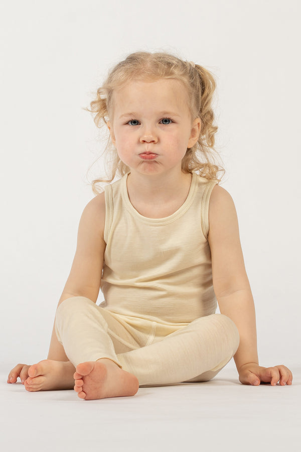 A young child with light curly hair in pigtails is sitting on a white surface against a plain white background. The child is wearing a menique sleeveless top and matching leggings. They are looking directly at the camera with a pouty expression, and their bare feet are visible.