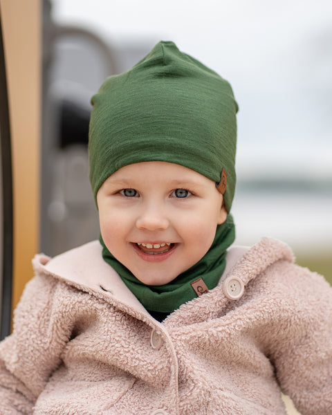 Happy toddler in dark green Merino wool kids’ beanie and matching neck gaiter, wearing a beige coat, smiling at the camera.