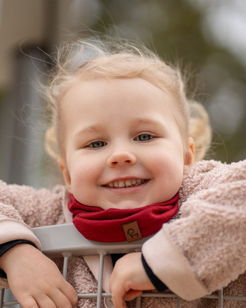 A smiling child with blonde pigtails and blue eyes looks happily at the camera, wearing a red Menique neck gaiter and a cozy beige jacket, leaning over a metal railing. The child&#39;s joyful expression fills the frame.