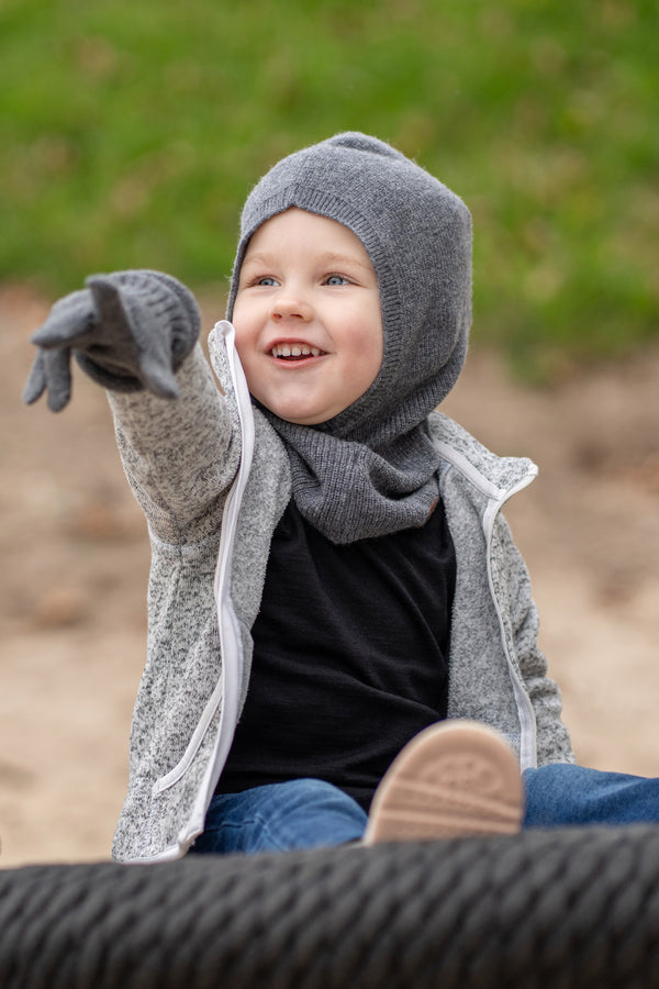 A young child with blue eyes and a wide smile, is sitting on a black rope swing, pointing to the left with their arm outstretched. They are wearing a dark grey balaclava, a grey speckled zip-up hoodie, dark grey mittens, blue jeans, and light pink sneakers. The background is a blurred outdoor scene with sand and grass.