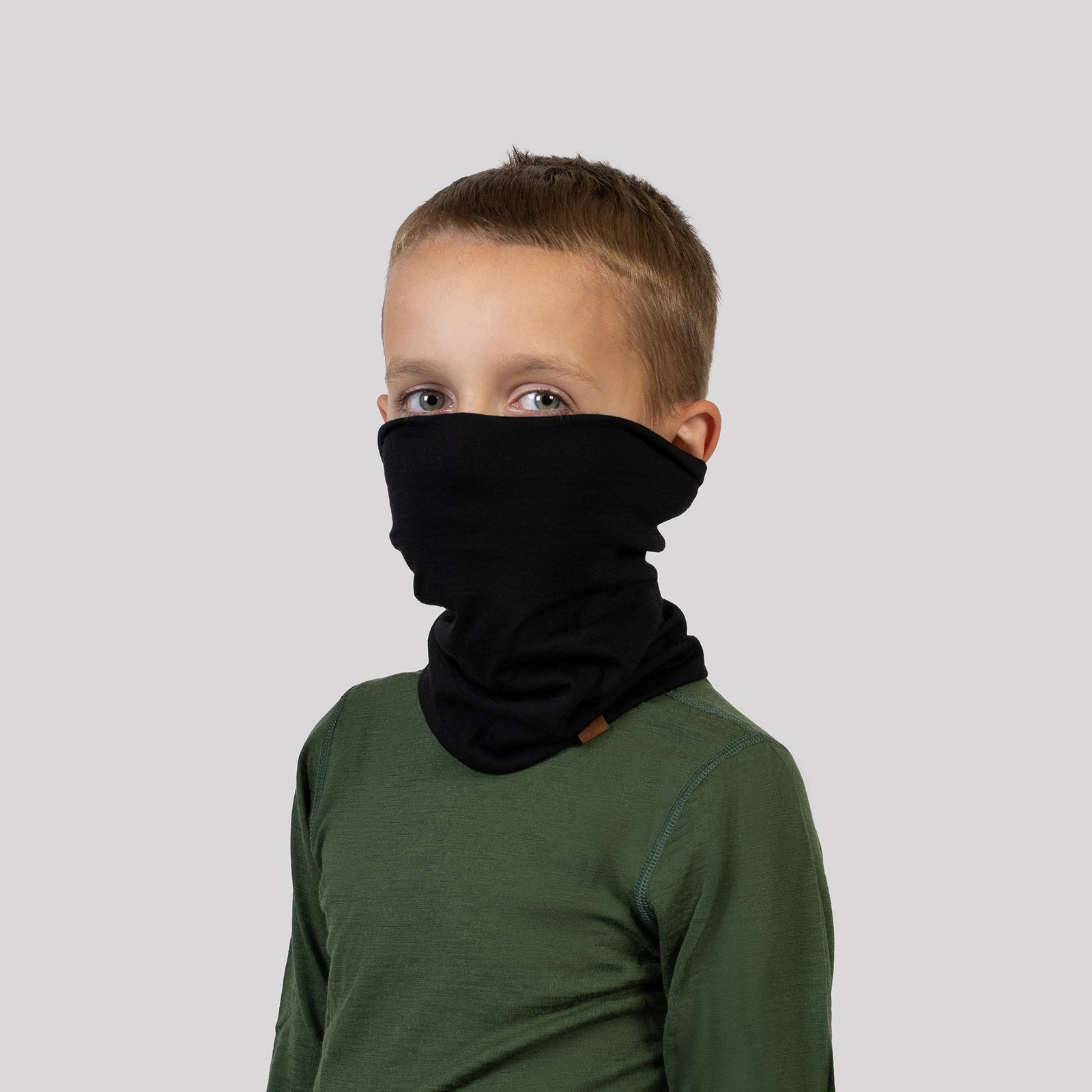 A cheerful child with blonde pigtails looks straight and to the left, peering through a yellow and gray circular opening on a playground. The child is wearing a Black Menique neck gaiter and a fluffy beige jacket, with a slight smile on their face.