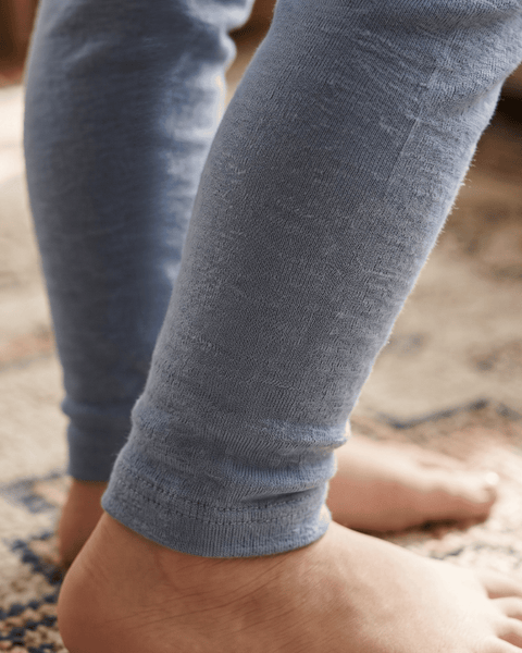 Close-up of a child's lower legs and bare feet standing on a patterned rug, wearing fine-knit, dusty blue leggings with cuffed ankles.