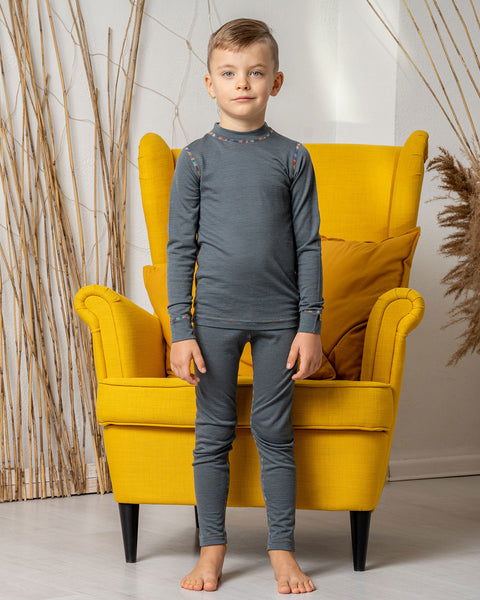 A boy standing in front of a yellow armchair, wearing the menique Kids&#39; 160 Merino Pants in Perfect Grey color with a matching long sleeve top. 