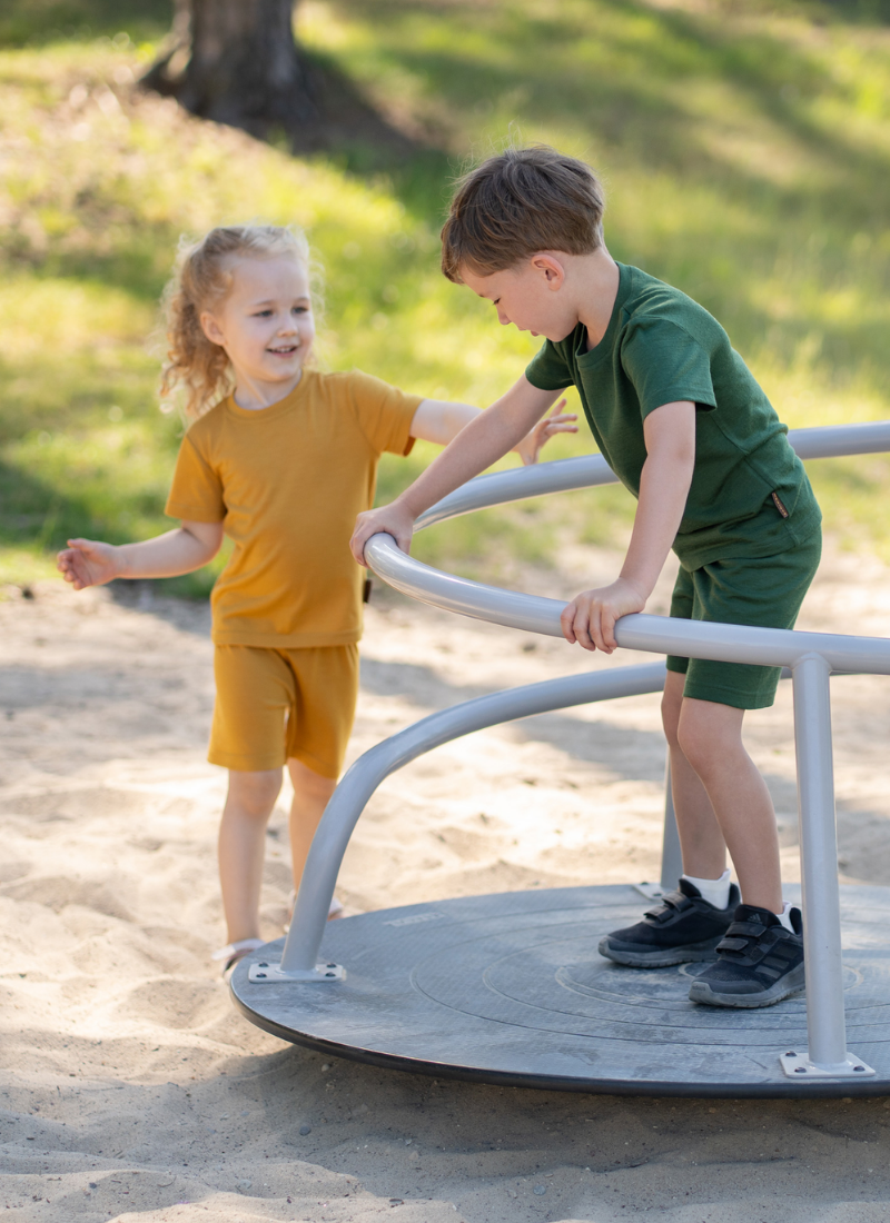 Two young children playing on a metal playground spinner in a sandy area. A boy standing on the spinner wears a forest green short-sleeve top and shorts set, while a girl standing beside it wears a mustard yellow short-sleeve top and shorts set.