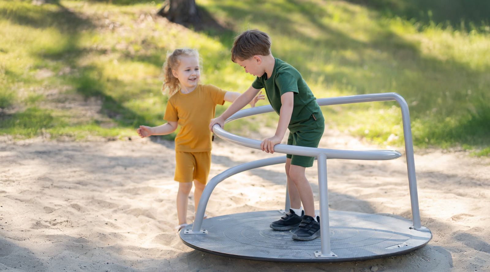 Two young children playing on a metal playground spinner in a sandy area. A boy standing on the spinner wears a forest green short-sleeve top and shorts set, while a girl standing beside it wears a mustard yellow short-sleeve top and shorts set.