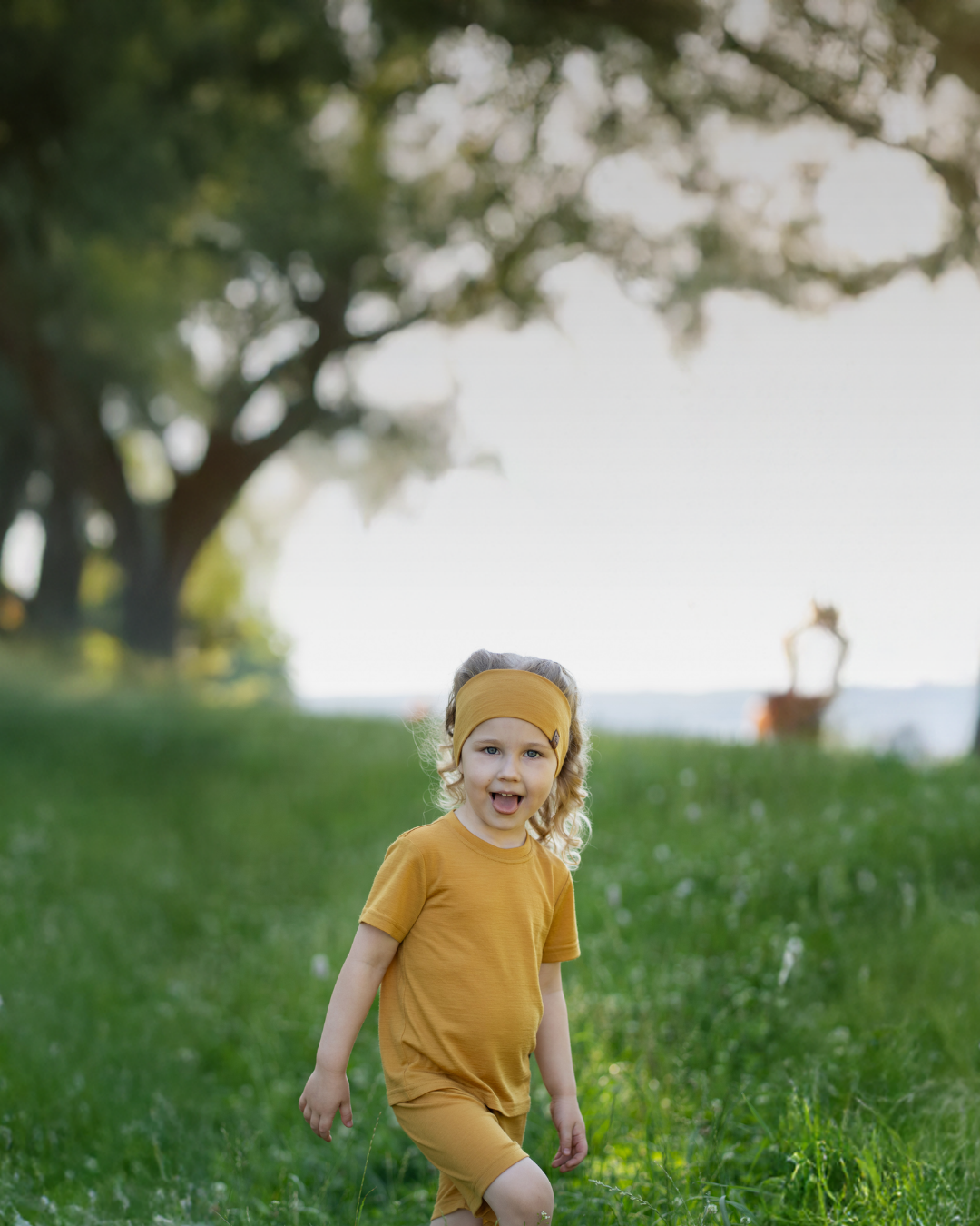 A young girl with curly blonde hair playfully sticking her tongue out, wearing a matching mustard yellow headband, short-sleeve t-shirt, and shorts while walking in a lush, green field.