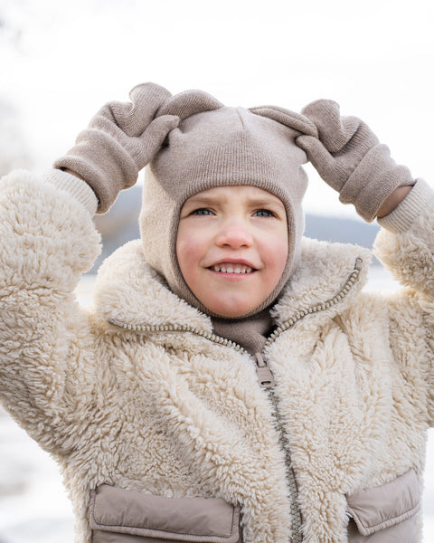 A child wearing a creamy beige balaclava with ear-like protrusions, matching light brown gloves, and a cream-colored, textured zippered jacket stands outdoors. The child is looking slightly upwards and to their left with a small smile, and their gloved hands are raised, holding the top of the balaclava. The background is blurred and light, suggesting a snowy or icy landscape.