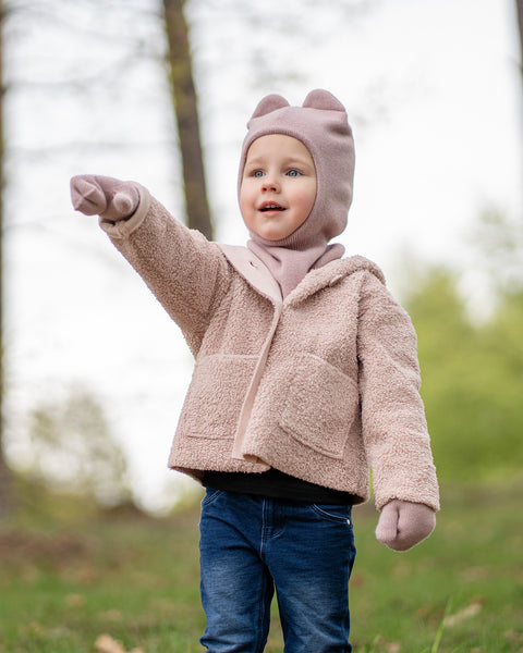 A young child is dressed in a menique dusty pink balaclava with ears, matching neck warmer and mittens, a fuzzy pink coat, and blue jeans. They are standing outdoors with their right arm extended, pointing, and looking intently towards the left. The background is a blurred natural setting with trees and greenery, possibly a park or forest.