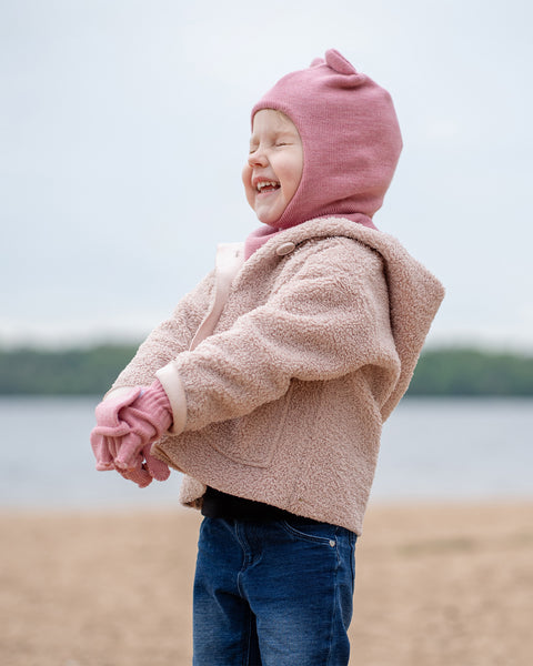 A joyful young child with eyes closed in laughter, stands on a sandy beach. They are wearing a pink balaclava with ears, a pink scarf, pink gloves, a beige sherpa coat, and blue jeans, with a blurred body of water and sky in the background.