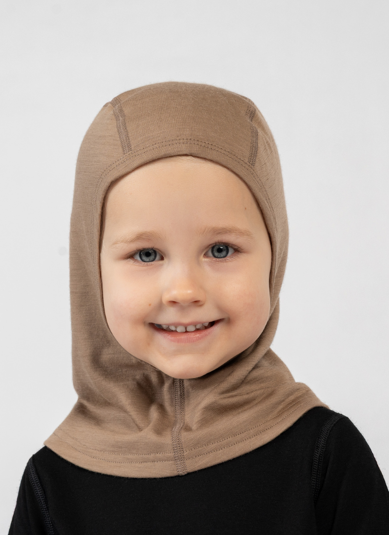 A young, smiling child standing against a white background. They are wearing a black long-sleeved top and a snug, beige balaclava that covers their head and neck while leaving their face exposed.
