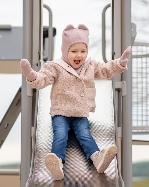 A young child with an open-mouthed expression of excitement, is captured mid-action sliding down a metal playground slide. They are wearing a pink balaclava with ears, a beige sherpa-style coat, blue jeans, and pink sneakers, with their arms outstretched.