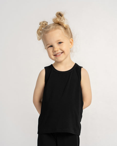 Smiling little girl in a black sleeveless top and leggings, standing barefoot with hands behind her back against a neutral background.