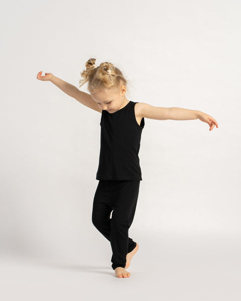 Child wearing a black sleeveless top and leggings set, balancing with arms outstretched on a light background.