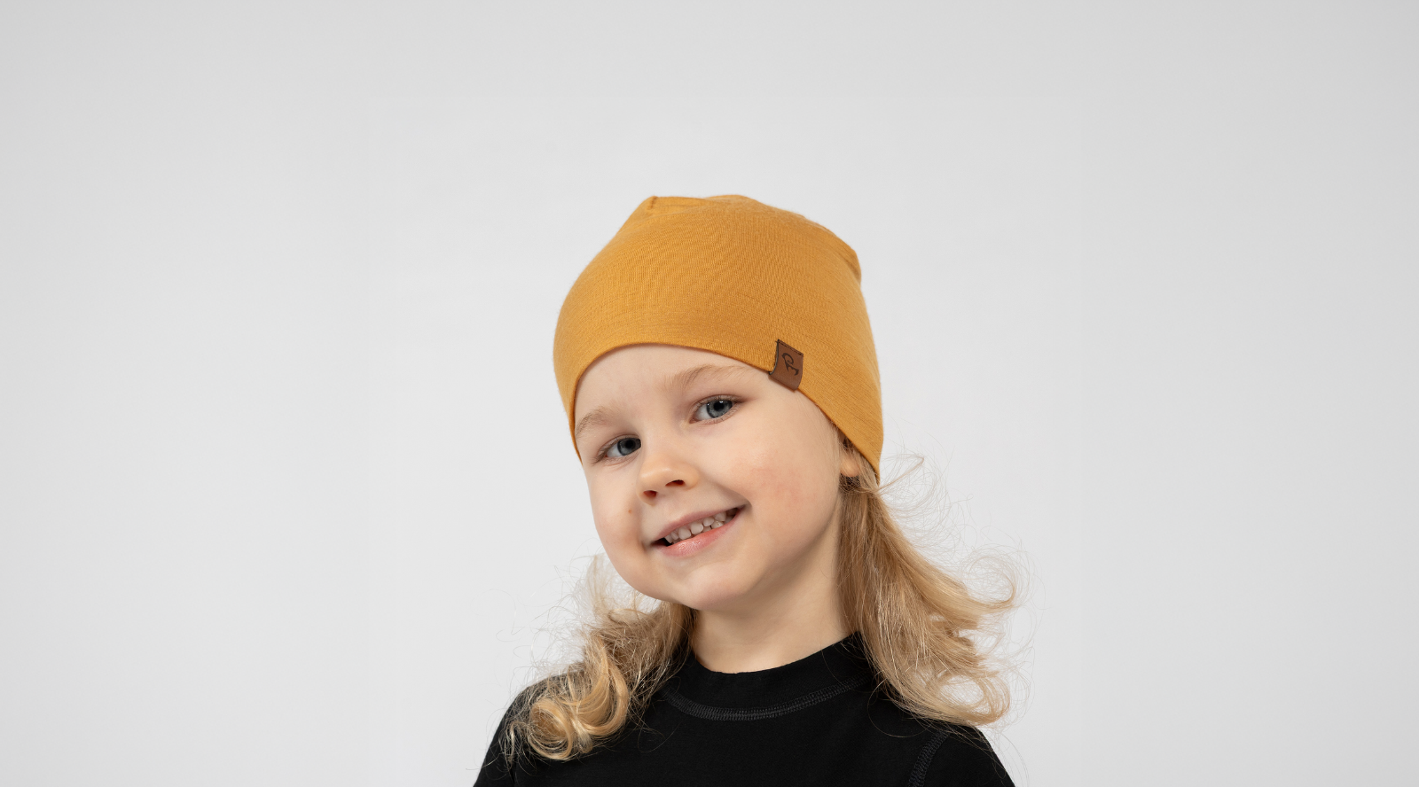A young child with blonde curly hair smiling brightly at the camera against a white background. They are wearing a black top and a mustard yellow beanie featuring a small brown logo tag on the edge.