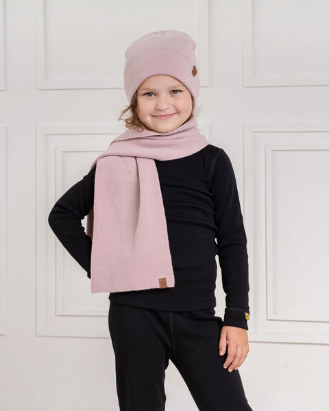 A child with wavy brown hair, wearing a dusty pink menique knit beanie with a small brown tag, a long pink knit scarf, a black long-sleeved shirt, and black pants, stands smiling at the camera. The child&#39;s right hand is on their hip, and their left arm is relaxed. The background is a white wall with decorative rectangular molding.