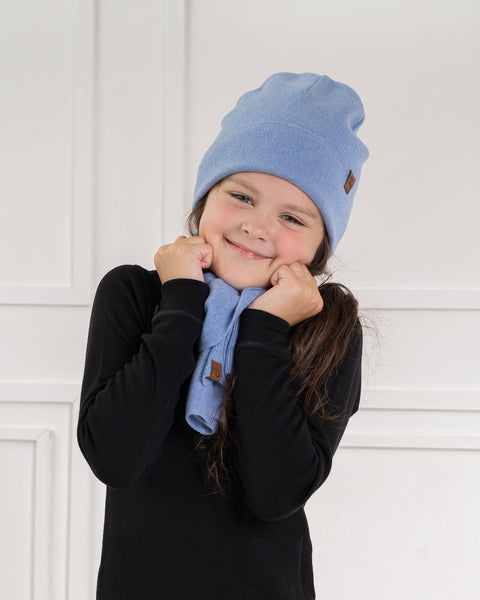 A smiling child with long, dark hair wears a light blue menique knit beanie with a small brown tag and a matching light blue knit scarf. They are wearing a black long-sleeved shirt and are holding their hands up to their cheeks, appearing playful. The background is a white wall with decorative molding.
