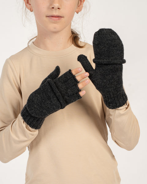 Girl wearing dark gray knitted convertible mittens, one flap closed and one open, beige top in neutral background.
