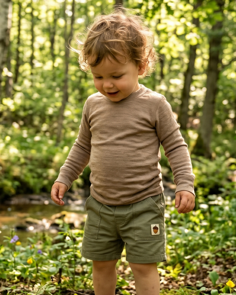 A smiling toddler stands outdoors in a lush, green forest setting, looking down gently at the ground. The child is wearing a light brown long-sleeve top paired with olive green shorts that feature a small acorn patch on the pocket.