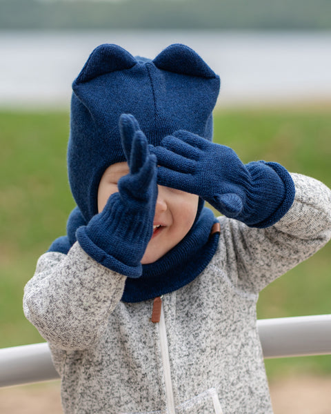 A young child, appearing to have light-toned skin, is seen from the chest up, playing outdoors. They are wearing a grey speckled zip-up jacket, a dark blue knitted menique balaclava-style hat with two rounded ears on top, and matching dark blue knitted gloves. The child&#39;s hands are raised to cover their eyes, as if playing peek-a-boo, but a wide smile is visible below their hands. A light-colored railing is in the foreground, and a blurred natural background with water is behind them.
