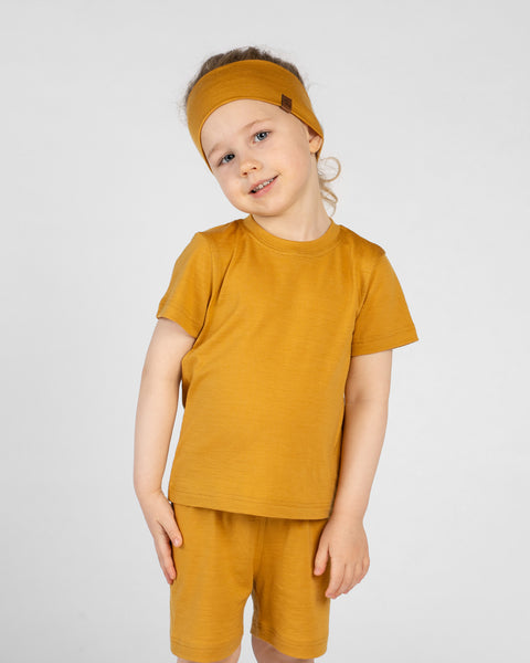 A child models a matching yellow set featuring a wide headband, a short-sleeve t-shirt, and shorts. They stand against a light gray background with a soft smile and their head tilted slightly to the side.