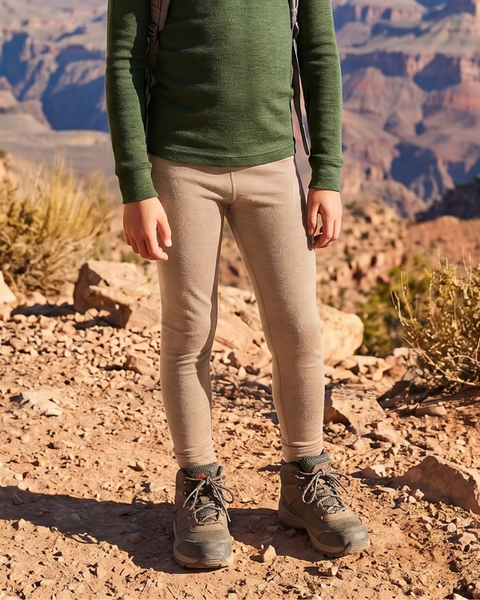 A close-up shot focuses on a child's legs and torso, standing on rocky terrain with a canyon in the background. The child is wearing a dark green long-sleeve top, beige leggings, and gray hiking boots.