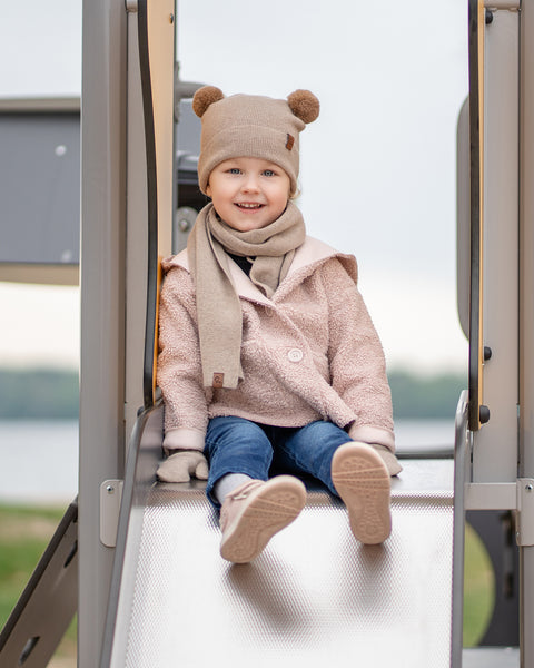 A young child with a cheerful smile, is sitting on a playground slide. They are dressed in a beige balaclava with brown pom-pom ears, a matching beige scarf, a light pink sherpa-style coat, blue jeans, and light pink sneakers. A blurred background shows water and trees.
