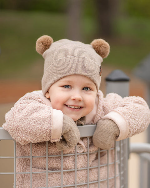 A young child with blue eyes and a wide smile, is looking directly at the viewer while peeking over a metal playground railing. They are wearing a beige knit hat with brown pom-pom ears, beige mittens, and a light pink sherpa-style coat.