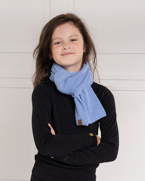 A young child with long dark brown hair, stands with a slight smile. They are wearing a black long-sleeved shirt, black pants, and a light blue knit scarf draped around their neck. The background is a white paneled wall.