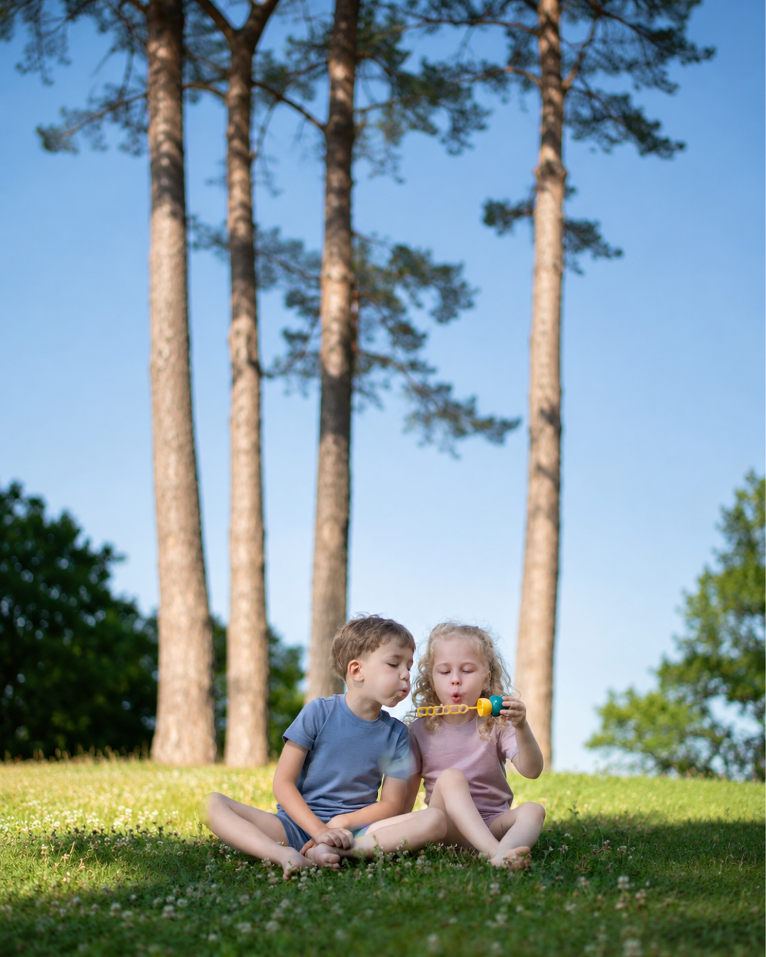 Two young children sitting on a grassy lawn outdoors, with tall pine trees and a blue sky in the background. The child on the right is blowing on a small yellow and blue toy.