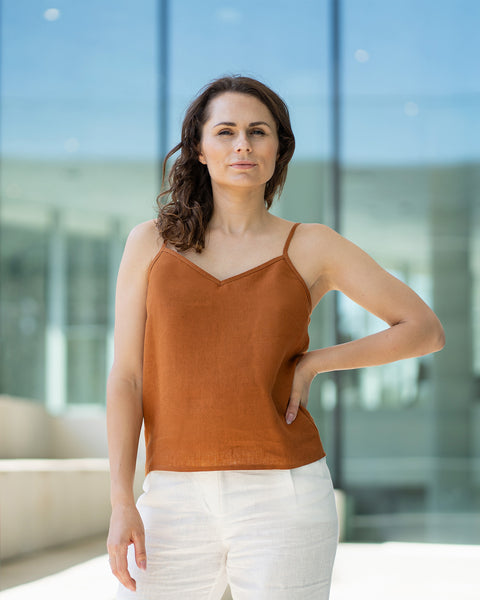 A woman with brown hair wears a almond brown V-neck camisole top and white trousers, posing indoors in front of large glass windows.